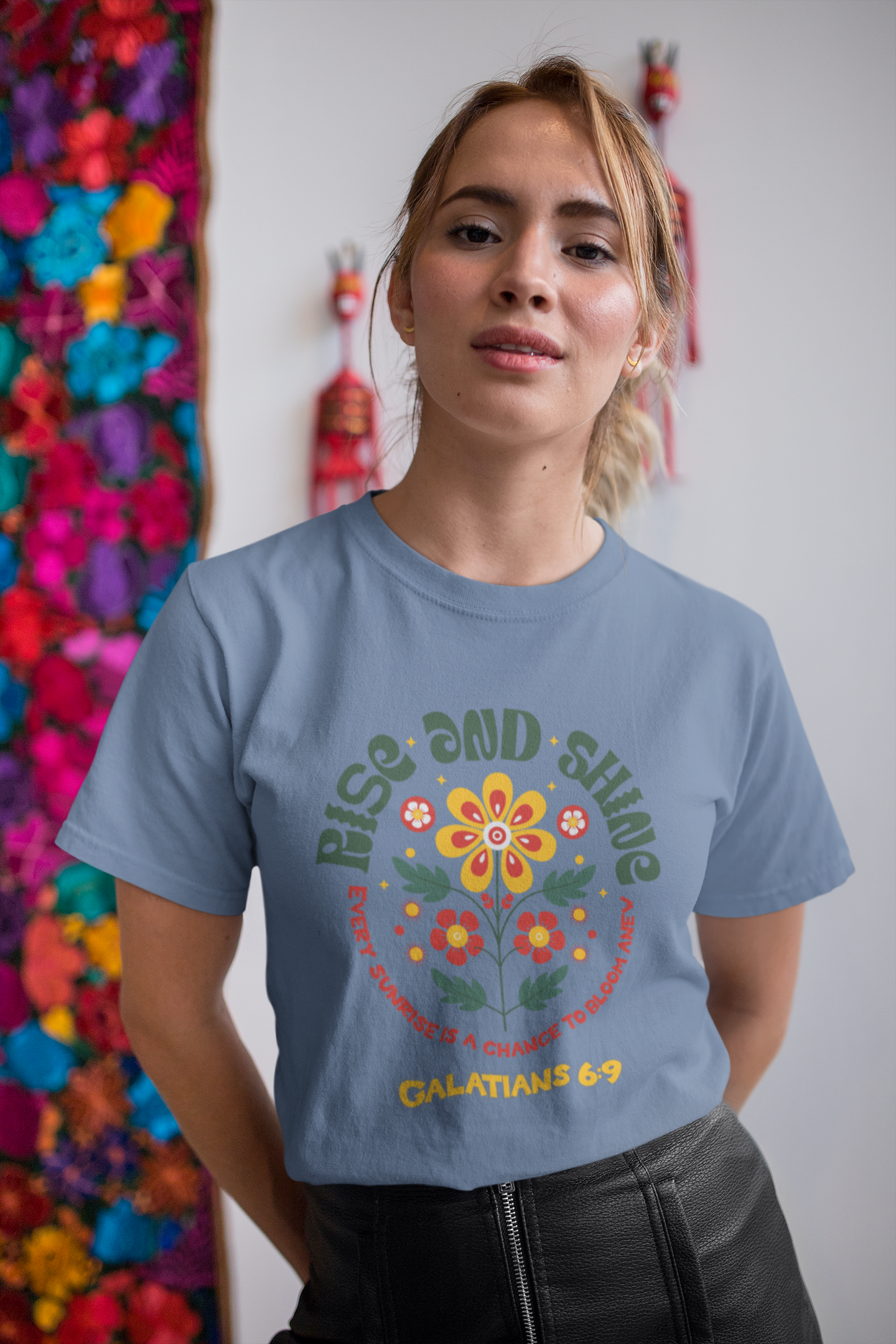 Woman wearing a blue t-shirt with a colorful floral design and text, standing in front of a wall with colorful flowers.